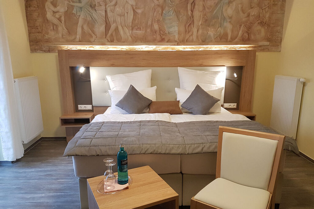 Modernes Doppelzimmer mit hellem Holzbetthaupt und klassischem Wandrelief, auf Beistelltisch Mineralwasser und zwei Gläser