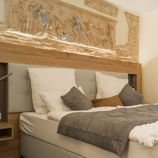 Schlafzimmer mit Doppelbett, weiß-grauer Bettwäsche, Zierkissen und beleuchtetem Friesrelief über dem Holzkopfteil