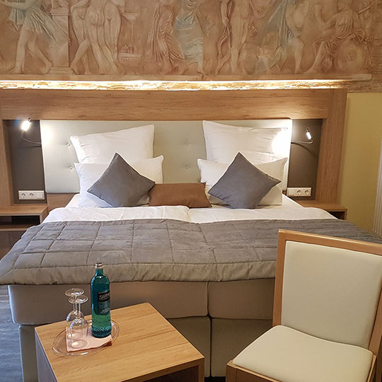 Gemütliches Hotelzimmer mit Doppelbett vor antikem Wandrelief und kleinem Tisch mit Wasserflasche und Gläsern