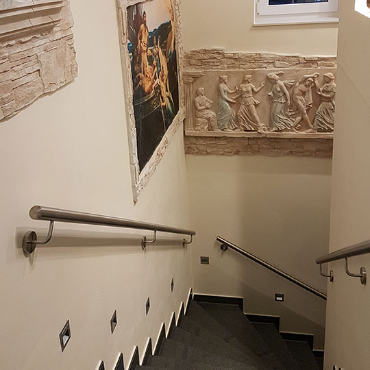 Innere Treppe mit grauem Teppich und Edelstahlhandläufen, Wände mit klassischem Relieffries und gerahmtem Mythologiebild