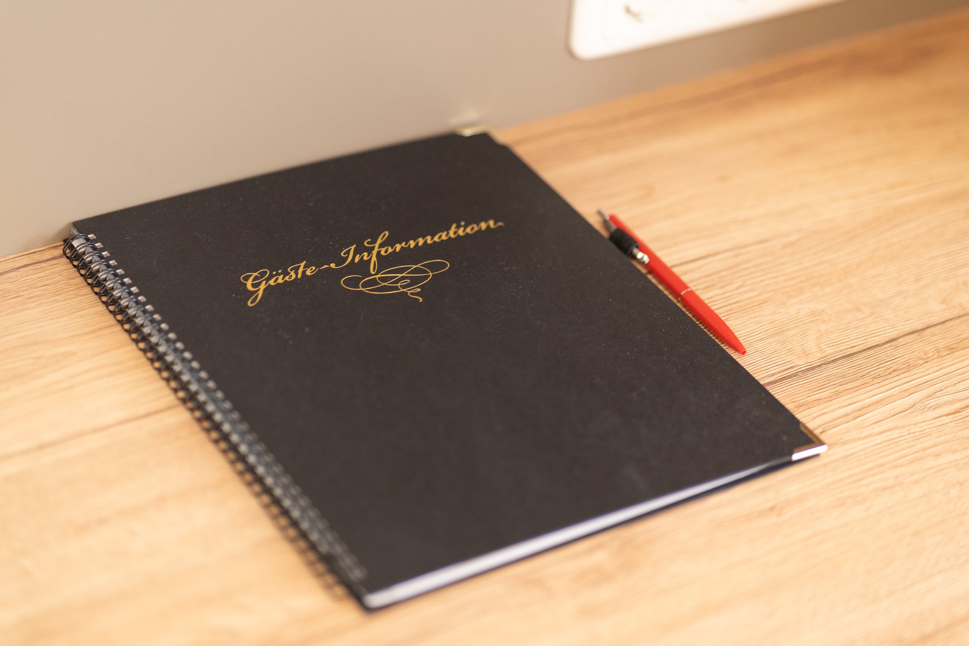 Schwarzes Gästeinformationsbuch mit goldener Aufschrift „Gäste-Information“ auf Holztisch neben rotem Stift
