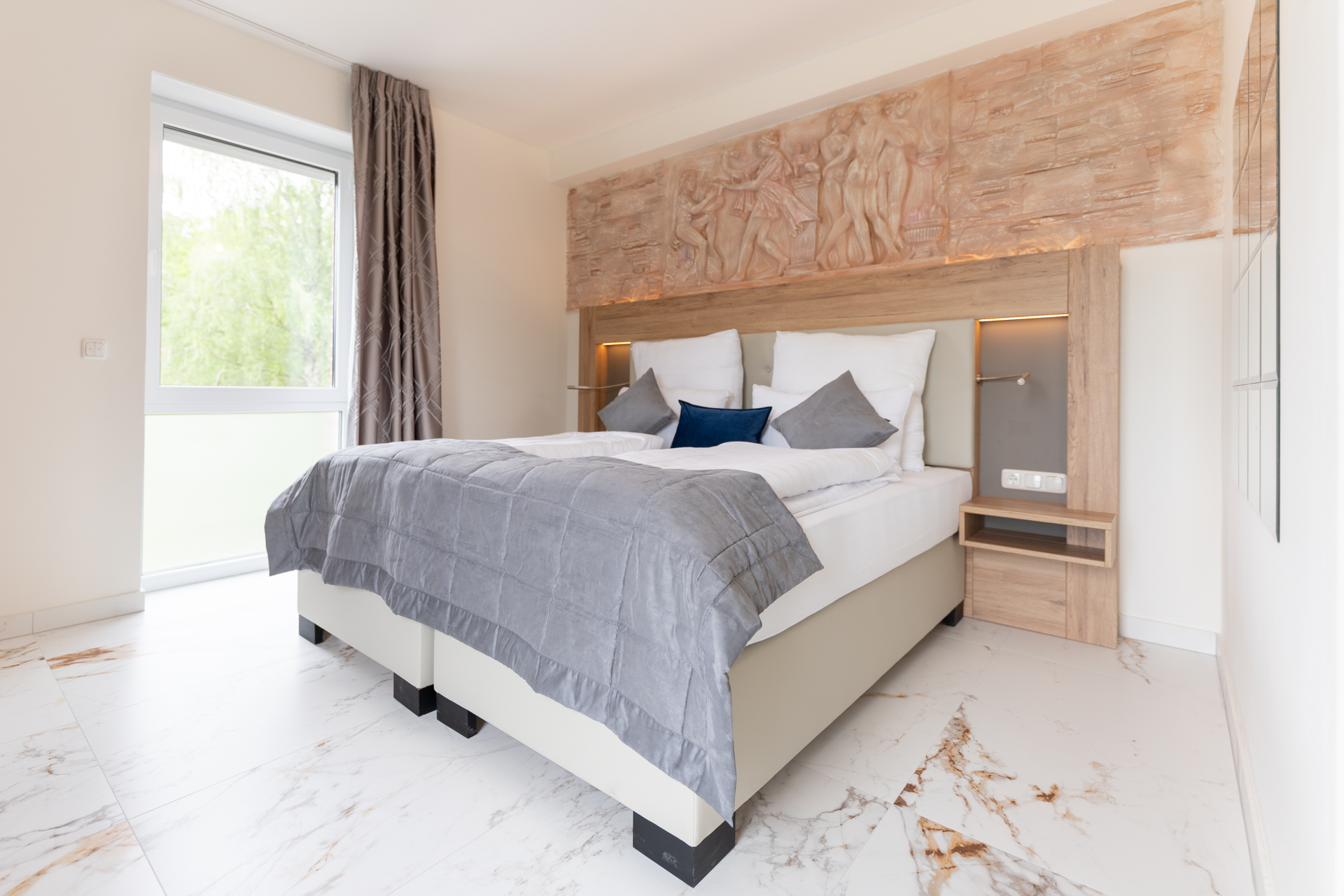 Modernes Schlafzimmer mit Doppelbett, grauer Bettdecke, weißen und blauen Kissen, hölzernem Kopfteil mit Reliefskulptur und Fensterfront