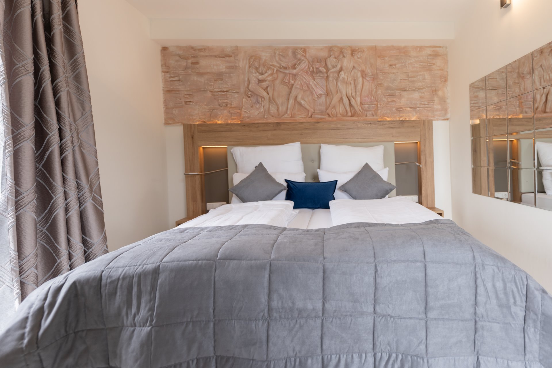 Doppelbett mit grauem Steppbett, weißen, grauen und blauem Zierkissen vor hölzernem Kopfteil mit antikem Relief
