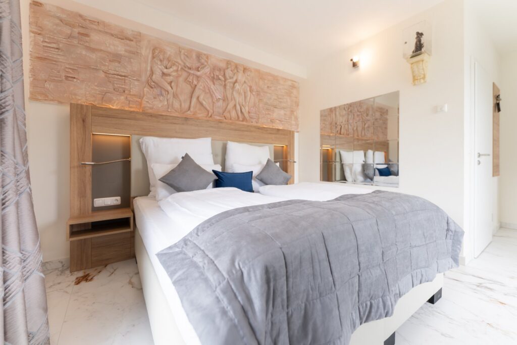 Helles Hotelzimmer mit weißem Doppelbett, grauer Tagesdecke, Holzkopfteil mit Figurenrelief, Spiegelfliesen und Marmorfußboden