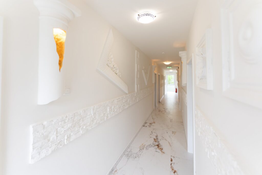 Heller Flur mit weißem Marmorfußboden, horizontalen Steinverblendern an den Wänden, weißen Reliefpaneelen und goldfarbenen Wandleuchten, am Ende ein Fensterlicht