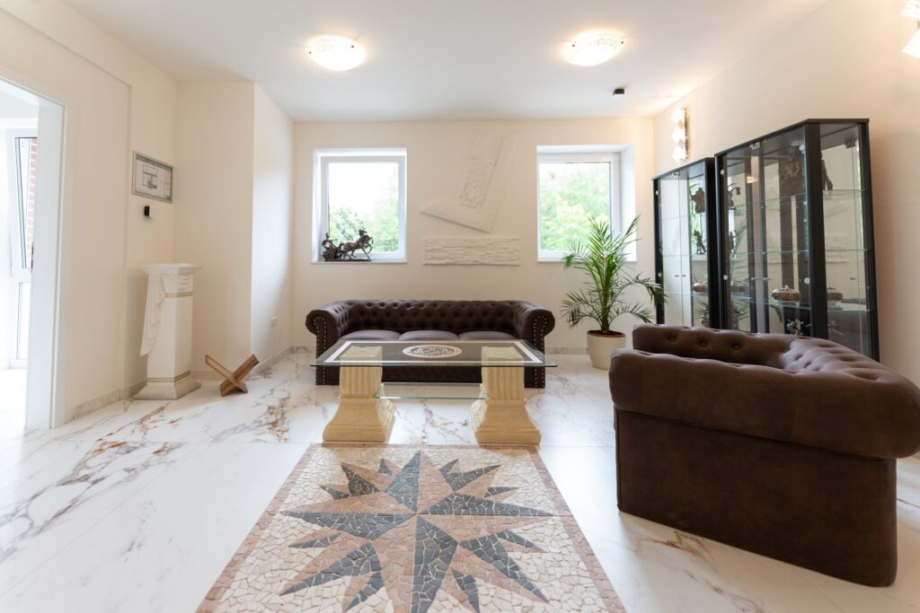 Helles Wohnzimmer mit zwei braunen Chesterfields, Glas-Couchtisch auf Steinsäulen, Mosaikboden und Vitrinen mit Skulpturen