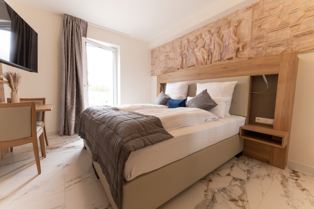 Modernes Schlafzimmer mit Doppelbett, grauer Steppdecke, Holz-Kopfteil mit mythologischem Relief und Schreibtisch am Fenster