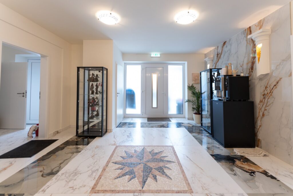 Heller Eingangsbereich mit Marmorboden und Kompassrosen-Mosaik, Vitrinen mit Statuetten, Kaffeemaschine auf Sideboard und Pflanze vor Glastür