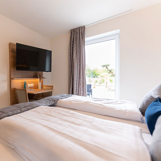 Hotelzimmer mit zwei zusammengeschobenen Betten, weißen Decken, grauem Läufer, Wand-TV, Schreibtisch und Terrassentür