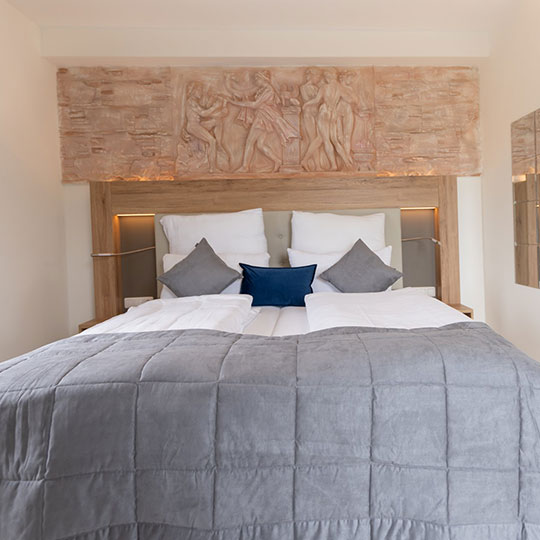 Doppelbett mit grauer Steppdecke, weißen Kissen und blau-grauen Zierkissen vor Holzkopfteil mit antikem Figurenrelief