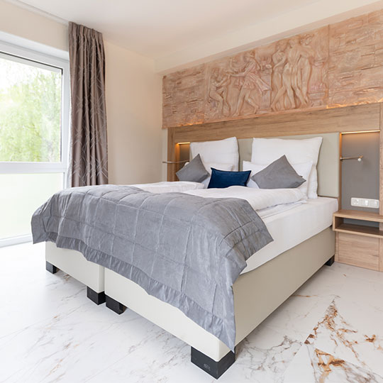 Doppelbett mit grauem Überwurf und Kissen in hellem Zimmer, Relief über dem Holz-Kopfteil und Fenster mit Vorhang