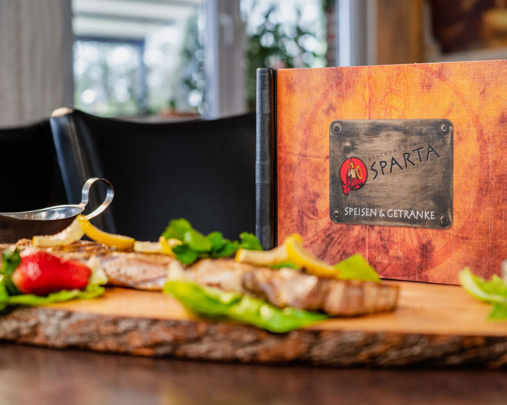 Steak mit Salat, Zitronenscheiben und Sauce auf Holzbrett neben der Speisen-und Getränkekarte des Restaurant-Café Sparta