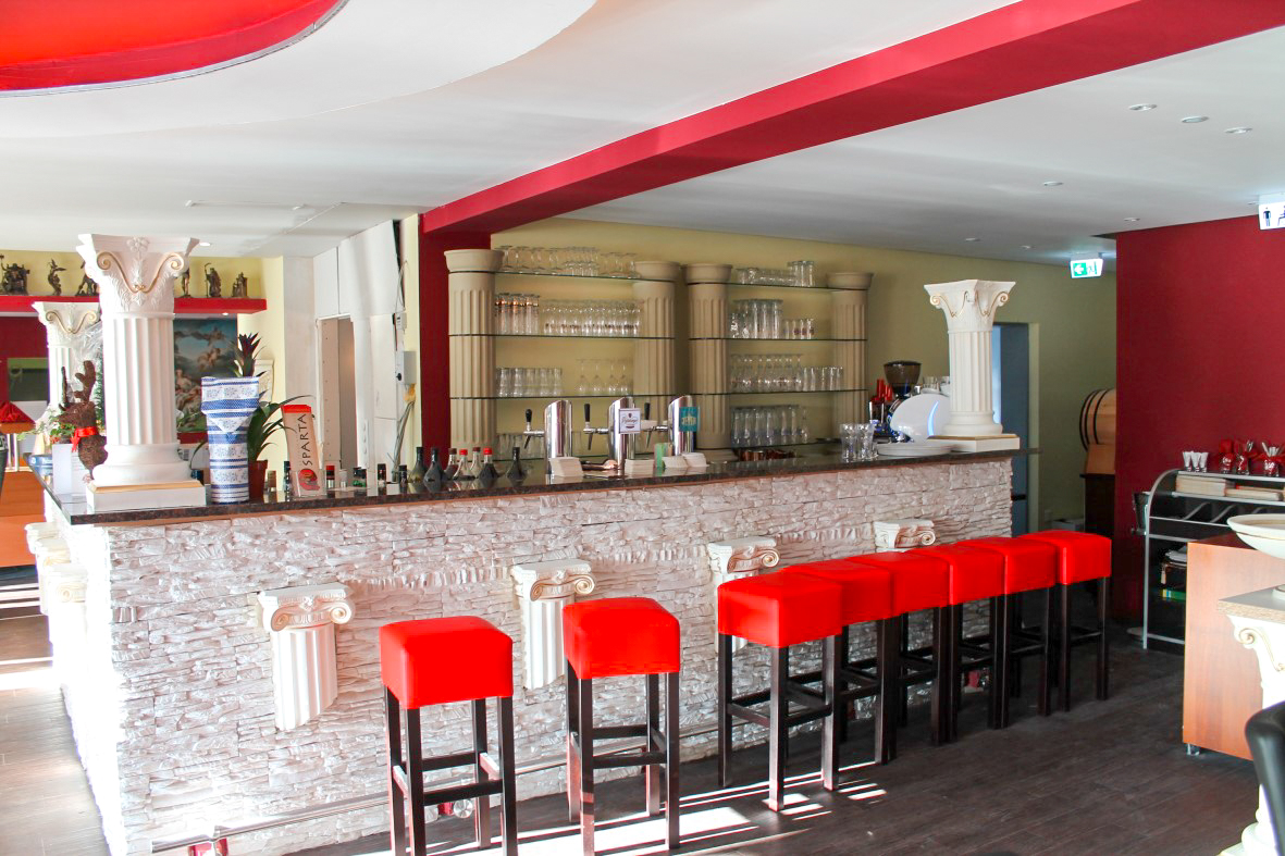 Bar mit weiß verkleideter Front, korinthischen Säulen und sechs roten Barhockern vor einem Regal voller Gläser