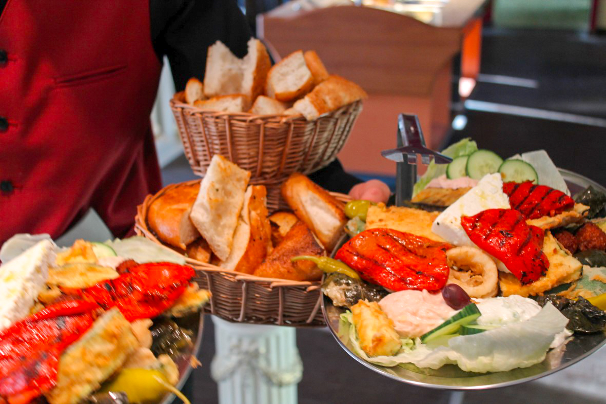 Kellner reicht Körbe mit Brot und zwei Mezze-Platten mit gegrillten Paprika, Dips, Feta und Gurkenscheiben