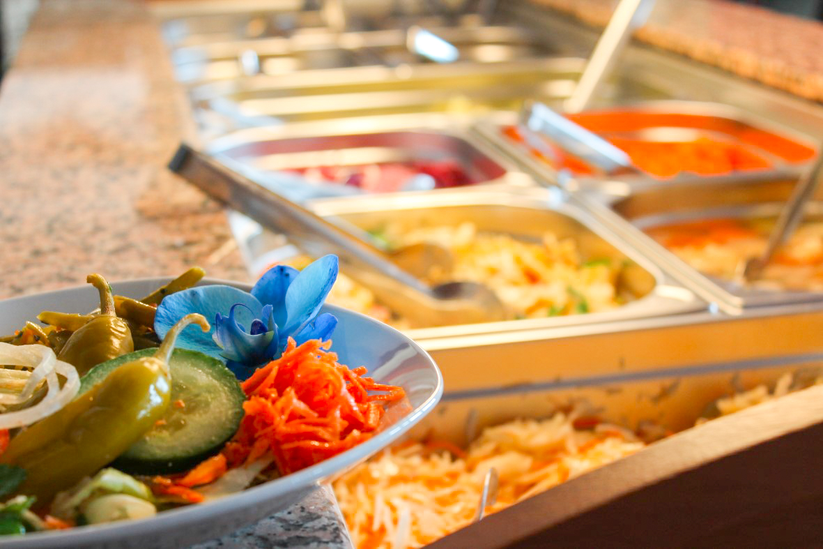 Schale mit eingelegten Chilis, Gurkenscheibe, geraspelten Karotten, Zwiebeln und einer blauen Orchidee vor einem Salatbuffet