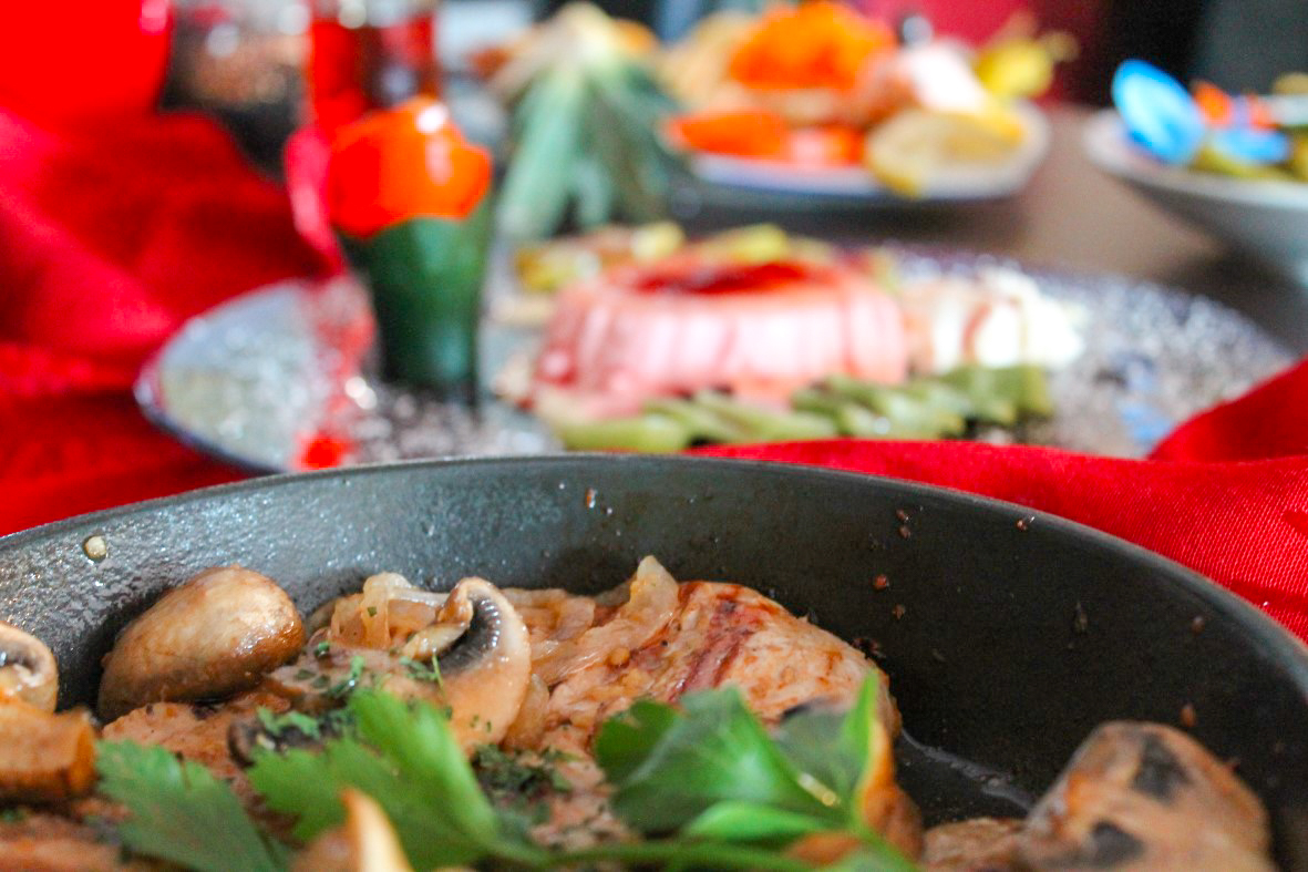 Pfanne mit gebratenem Fleisch, Champignons und Petersilie auf rotem Tuch, im Hintergrund verschwommene Vorspeisenteller