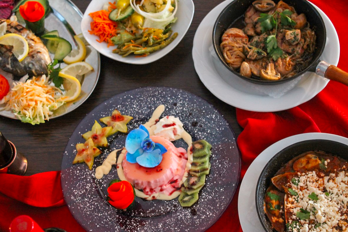 Rosa Dessertmousse mit blauer Orchidee, Sternfrucht und Kiwi auf schwarzer Platte, umgeben von Fisch, Salat, Pfannengerichten