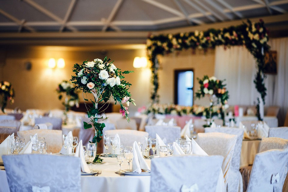 Festlich gedeckte runde Tische mit weißen Tischdecken und hohen Rosen- und Grünblumengestecken in einem Hochzeitsempfangsraum