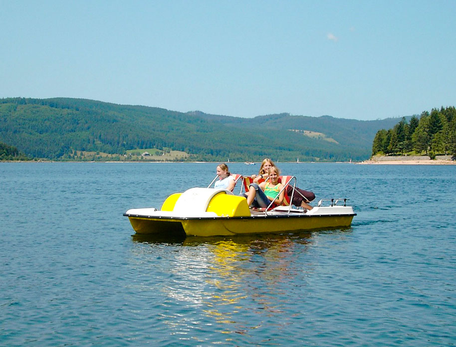 Drei junge Frauen sitzen in einem gelben Tretboot auf einem ruhigen Bergsee, umgeben von bewaldeten Hügeln