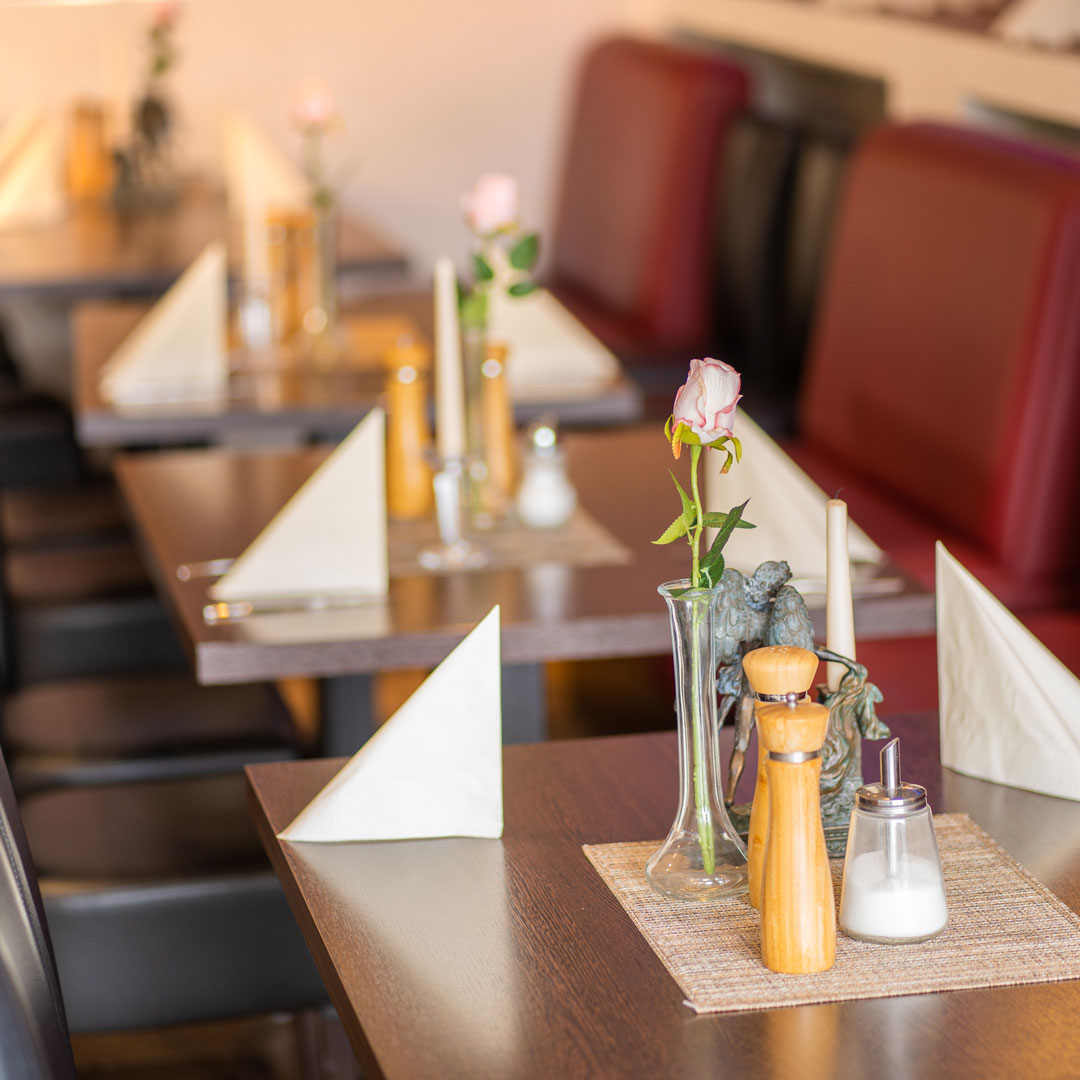 Reihenweise gedeckte Restauranttische mit aufgestellten weißen Servietten, einzelnen Rosen in schlanken Glasvasen, Pfeffer- und Salzstreuern sowie Zuckerdose
